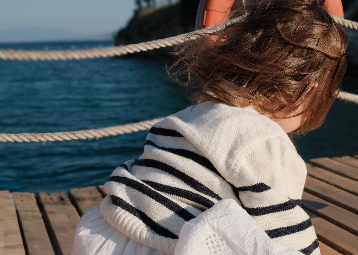 Child sitting on a wooden deck by the sea, wearing a striped sweater.